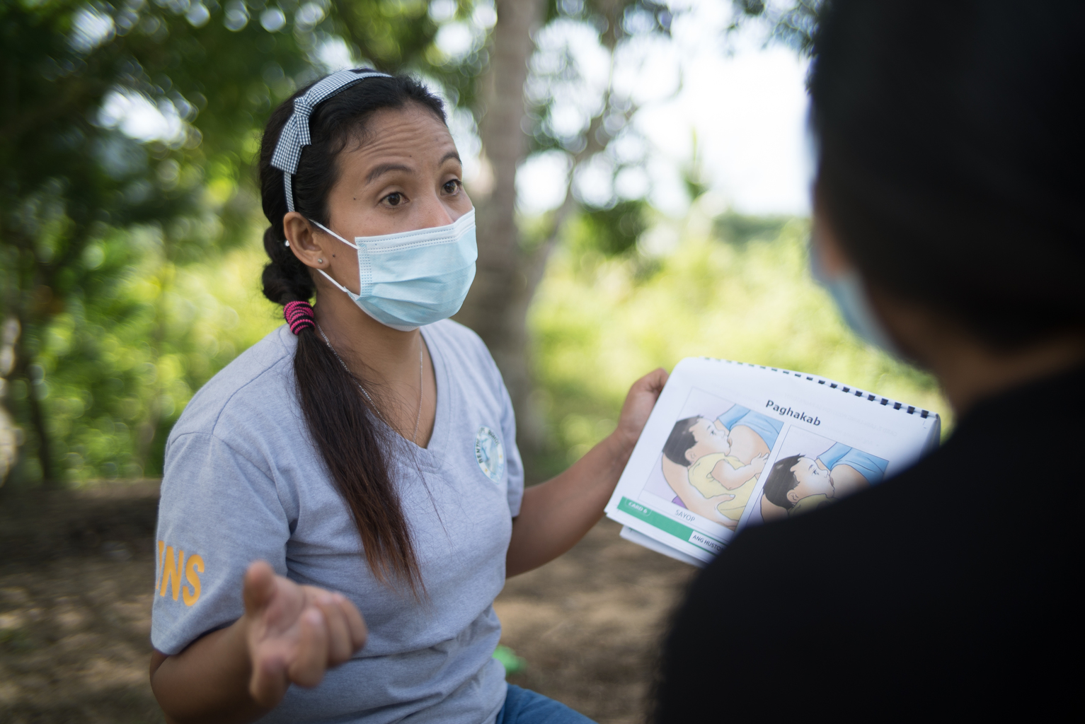 Philippines_BF1KD_DSC08337 - Nutrition International Woman wears a mask outside and holds a booklet during a mother-to-mother support group session in the Philippines