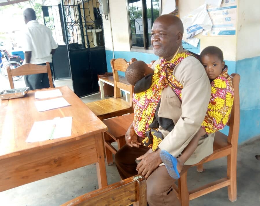 Tanzania_Lagangabilili_VAS_Alphonce Maduhu with son and daughter - Nutrition International Father sits on a chair at a health clinic carrying his son on his front and holding his daughter behind him.