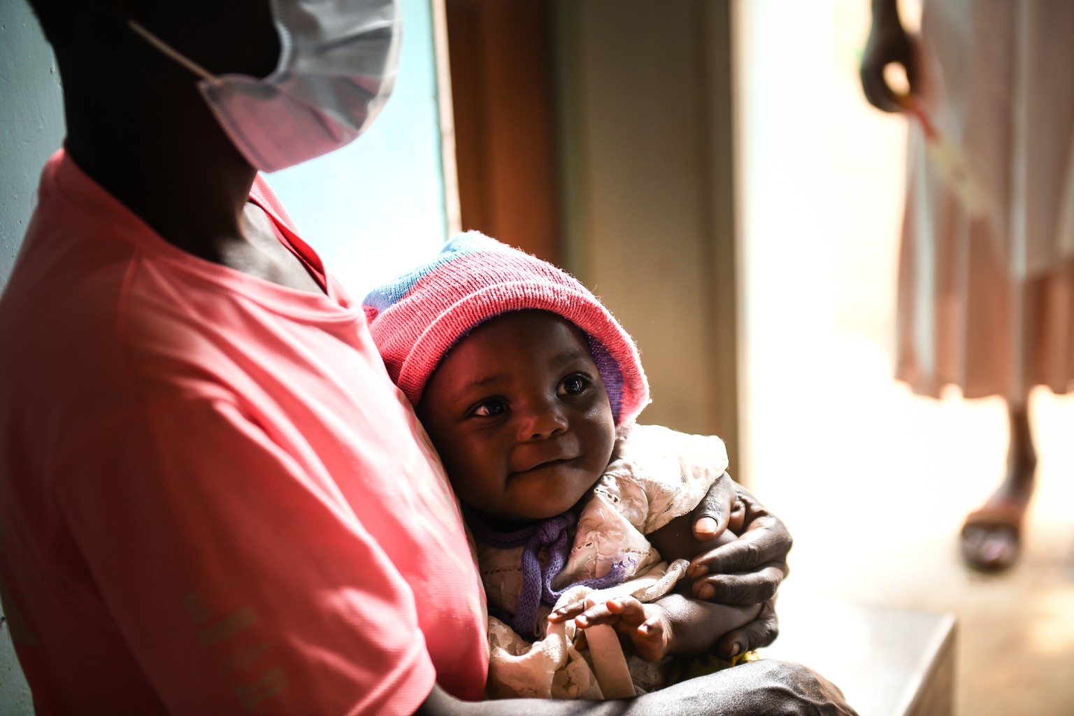 - Nutrition International An infant wearing a hat smiling in the arms of their mother as they wait to receive a vitamin A dose at Chisitu Health Centre in Mulanje southern Malawi.