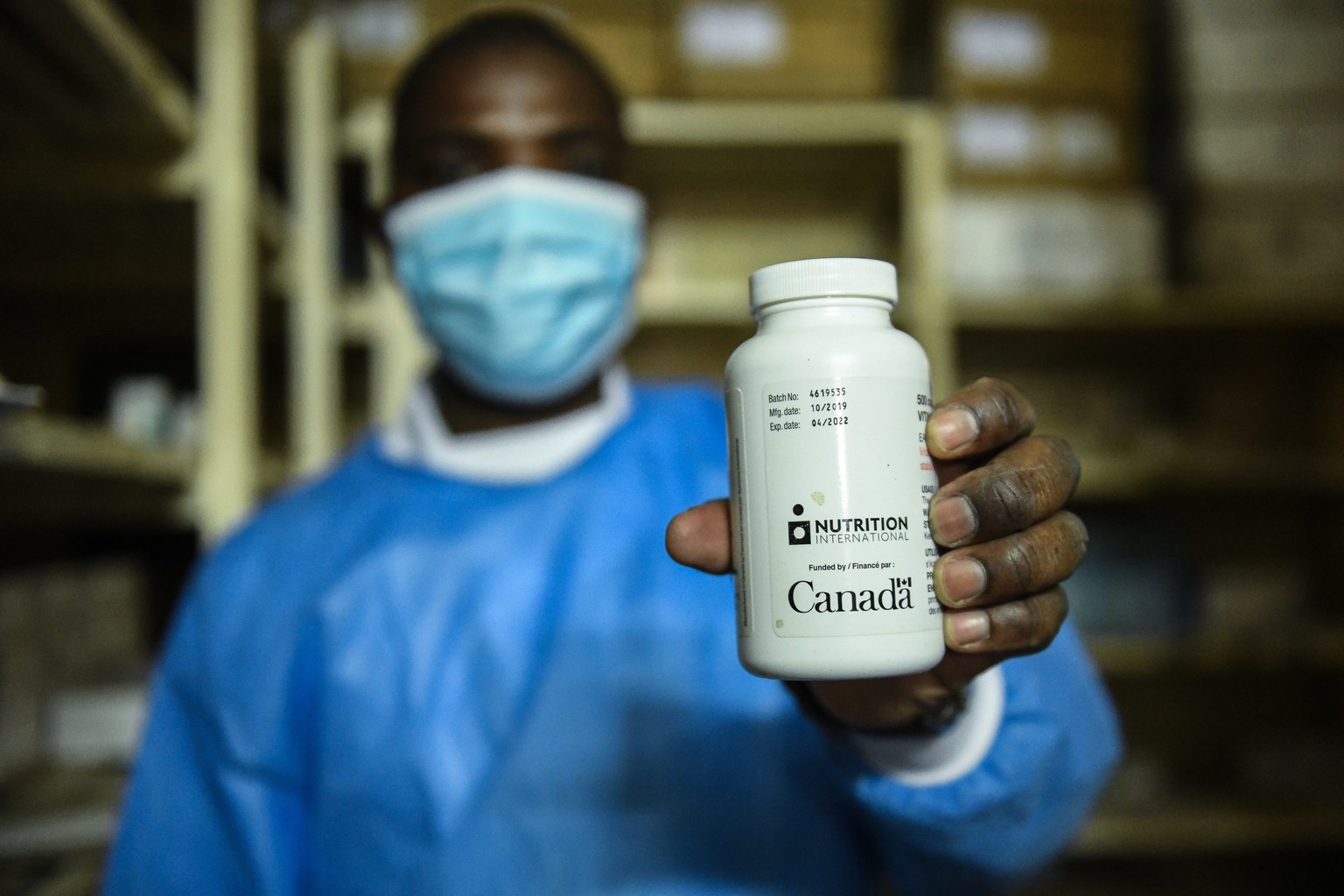 A health care working wearing scrubs and a mask holds up a bottle of vitamin A tablets
