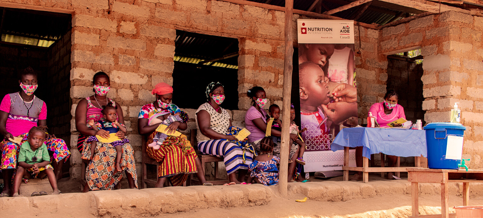 Mothers wait outside a health post on wooden chairs with their children in their laps as the nurse completes paperwork to prepare their children for a vitamin A dose.