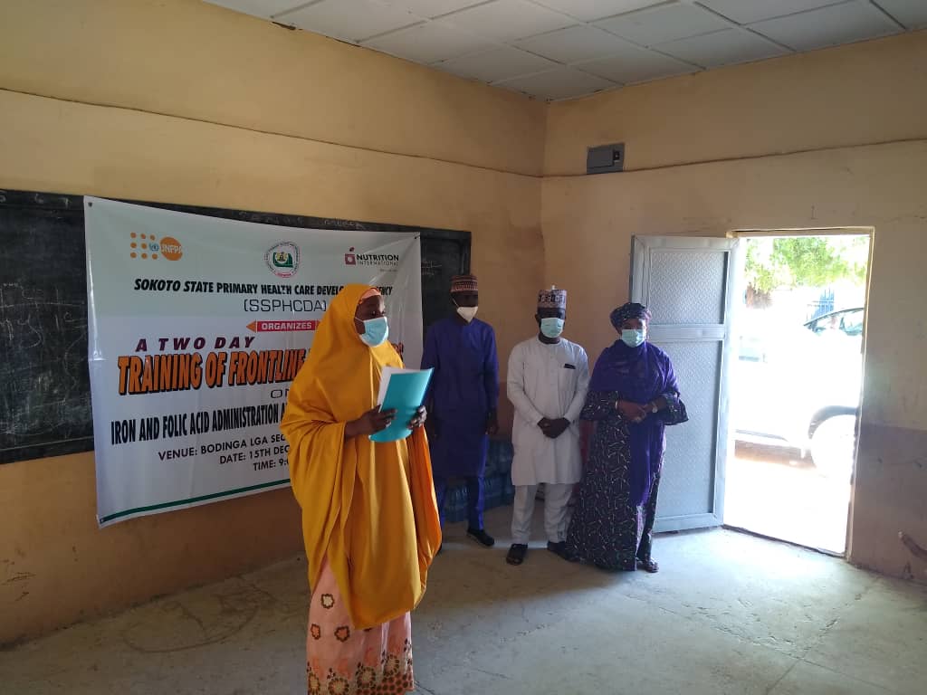 Nigeria_Sokoto_gender equality training_44638898 - Nutrition International Une femme se tient à l’avant d’une salle, avec en main une brochure qu’elle présente à l’auditoire.