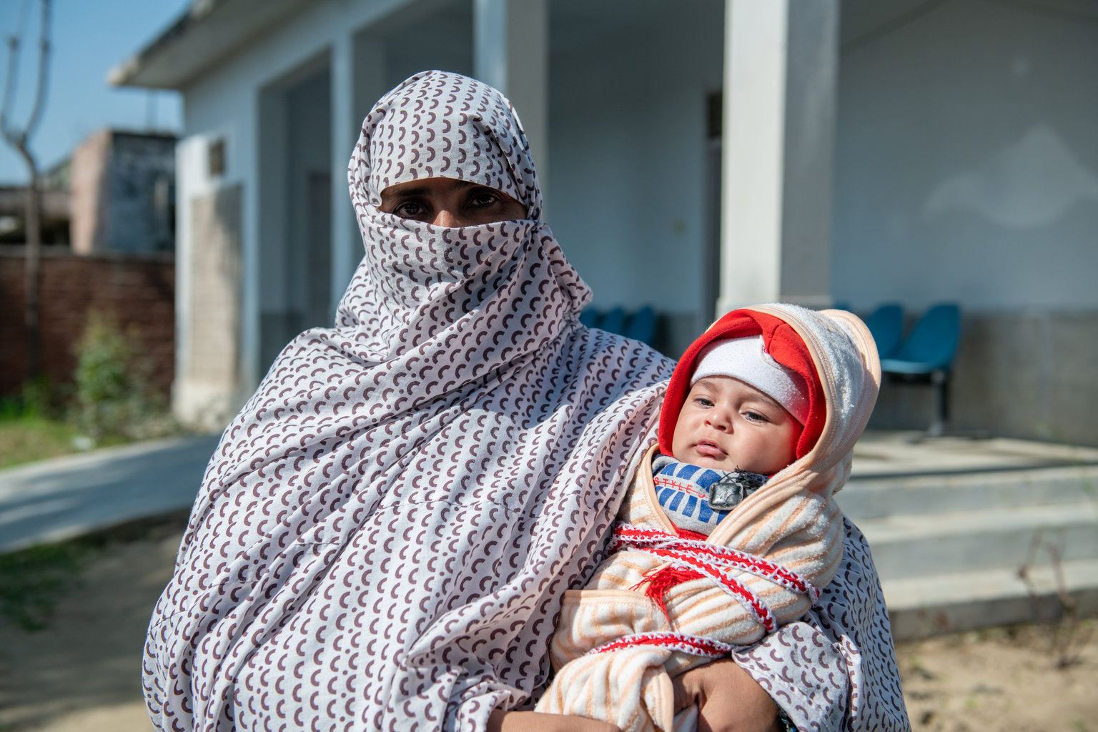 A woman looks at the camera wearing a white and red niqab and holding a young child in her arms, who is warmly wrapped in a blanket.<br />