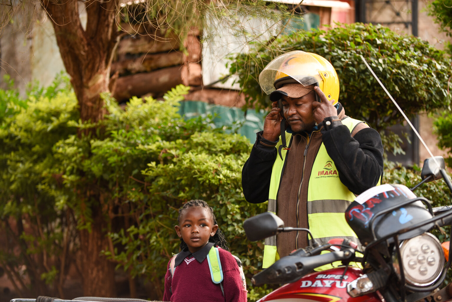 A man puts on a motorcycle helmet as he is preparing to leave for work. He is standing beside a young girl in a school uniform who is also leaving home for the day.