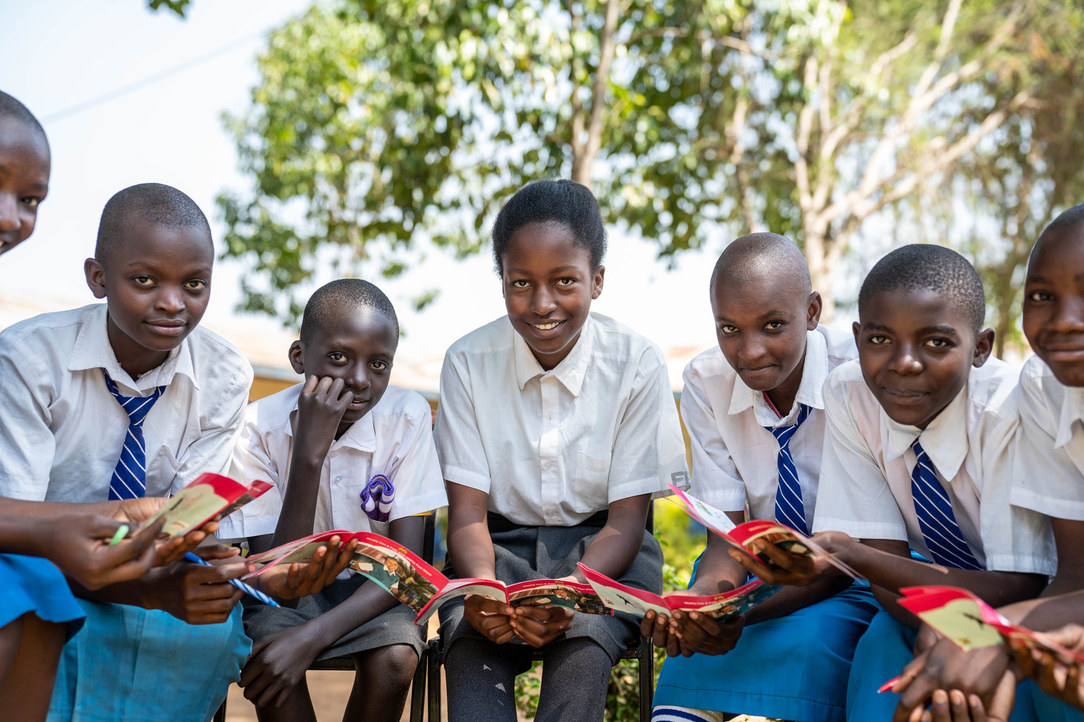 A group of students in school uniforms look up from reading pamphlets and smile at the camera. They are outside.