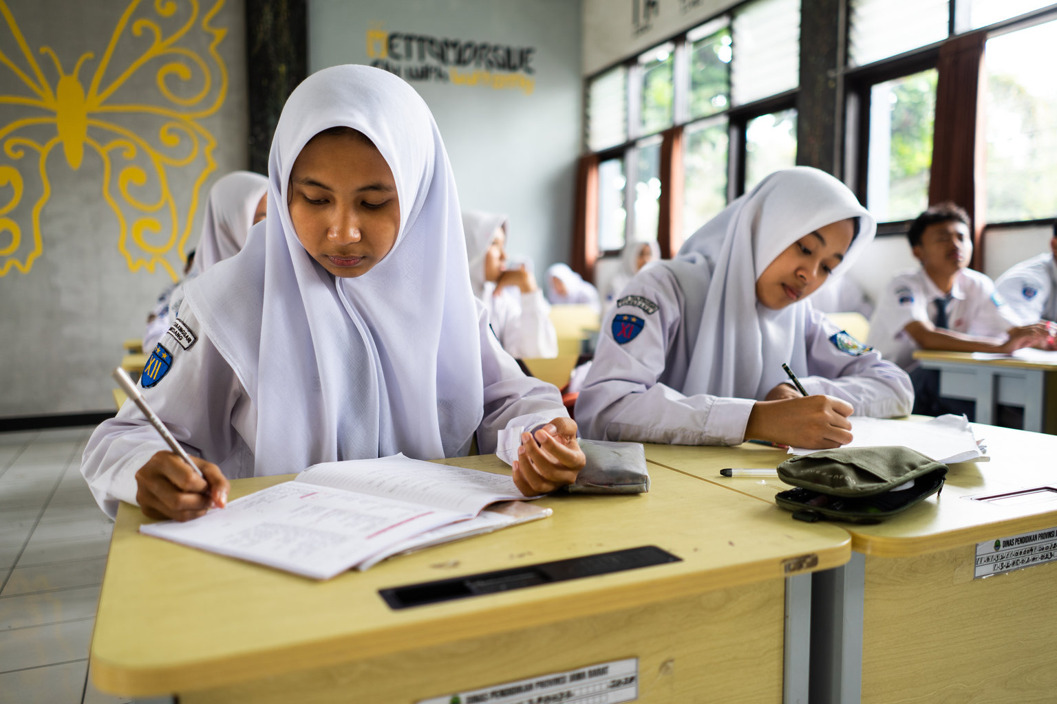 A girl sits at her desk in a classroom and writes into her notebook. She is sitting beside other female students who are also writing in their notebooks.