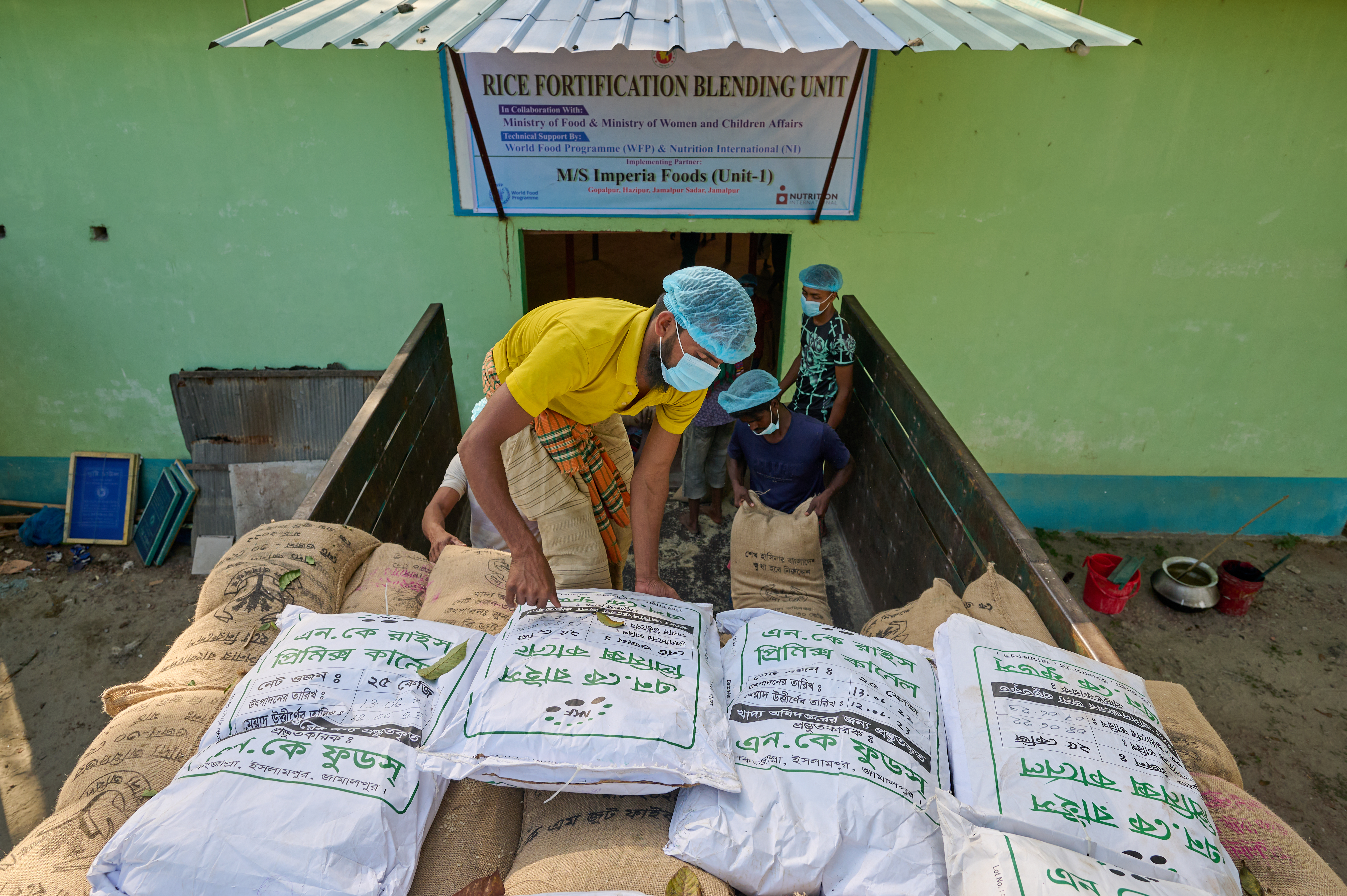 0078-NI-BD-20-11-2022-sujanmap - Nutrition International A rice fortification blending facility combines fortified rice kernels with non-fortified rice in the regulated ratio. Here, the rice is arriving on the back of a truck to be blended. Workers are unloaded the sacks of rice and carrying them into then blending facility.
