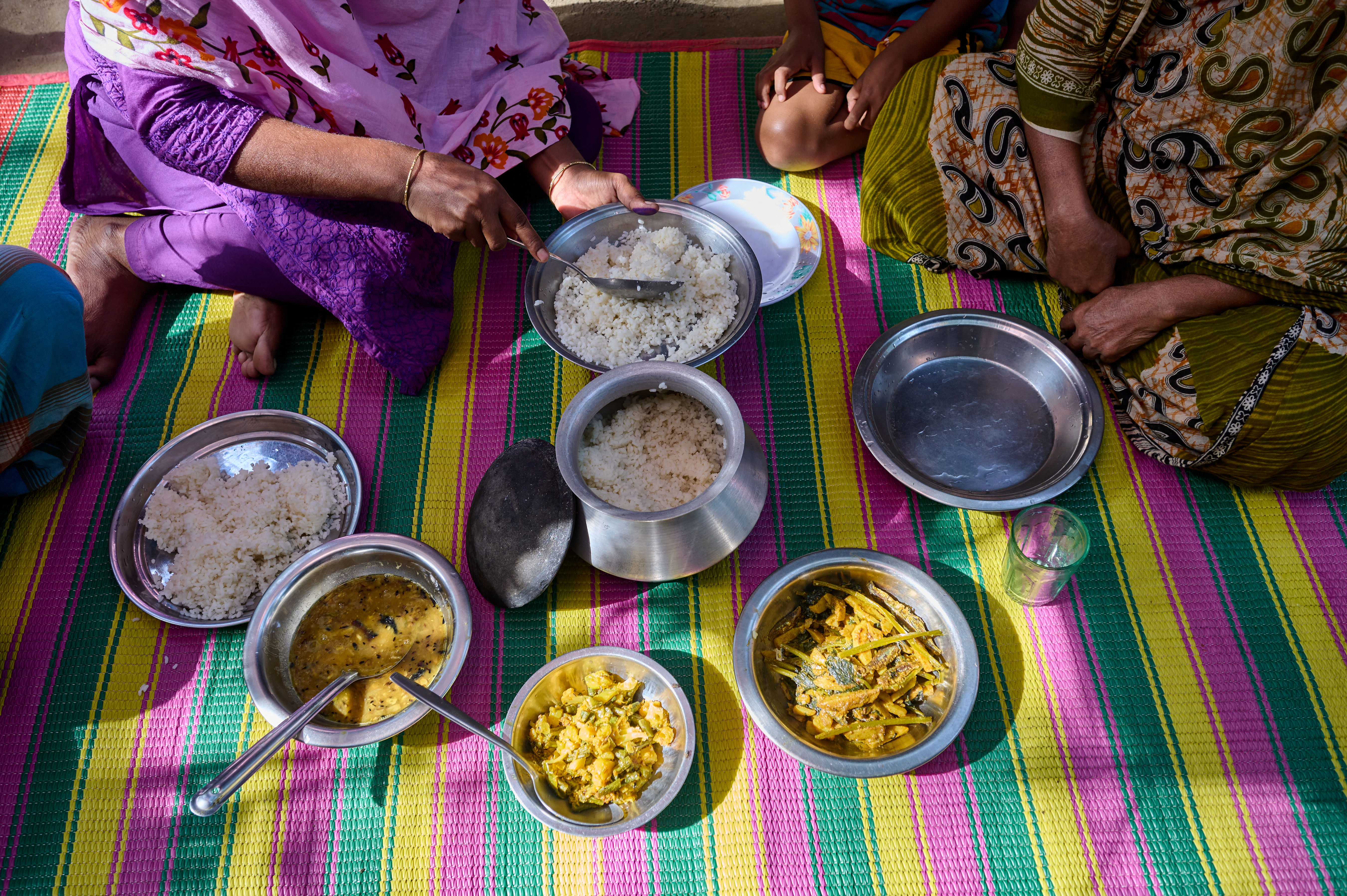 0133-NI-BD-21-11-2022-sujanmap - Nutrition International A midday meal is laid out on a colourful mat. Bowls of rice, and differnt kinds of vegetable curry are present. Two women sit cross legged on the floor, as one woman starts to serve the rice.