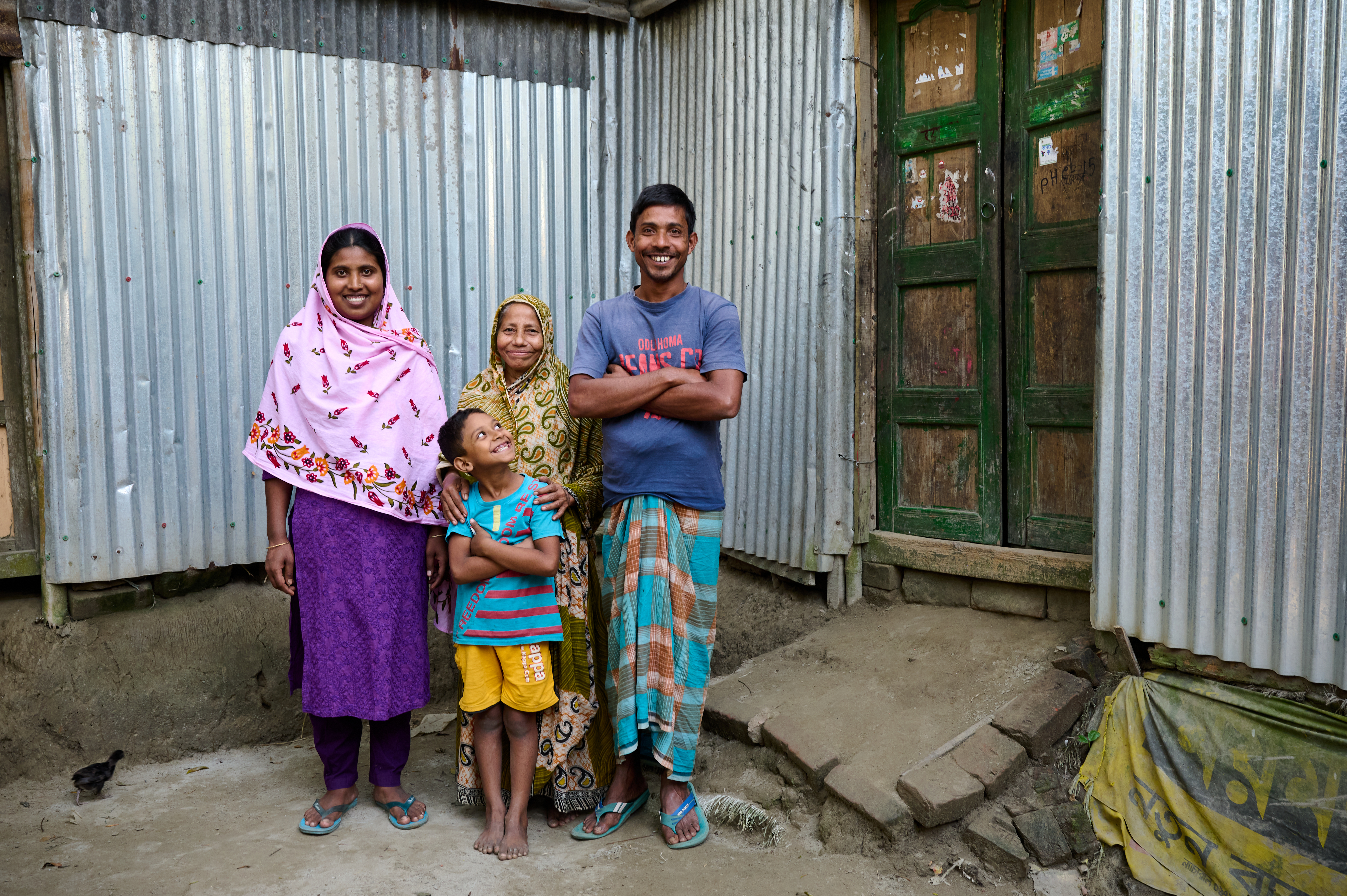 0152-NI-BD-21-11-2022-sujanmap - Nutrition International A woman stands with her mother-in-law, husband and child. All four are smiling and posing for the camera.